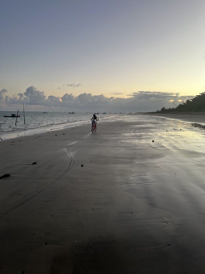 Mulher andando de bicicleta na areia molhada da Praia de Paripueira durante o pôr do sol, com mar calmo e céu alaranjado ao fundo.